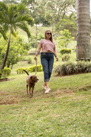 Vertical shot of a happy woman walking on the grass with her pet beside her outdoors. The woman is wearing jeans, sneakers, and sunglasses, and holding her dog on a leash