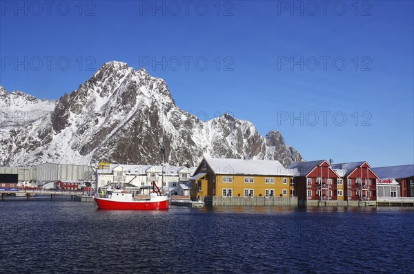 Colourful houses and a red fishing boat in the harbour in front of snow-covered mountains under a clear blue sky, Winter, Hotels, Svolvaer, Svinoya, Lofoten, Nordland, Norway, Europe