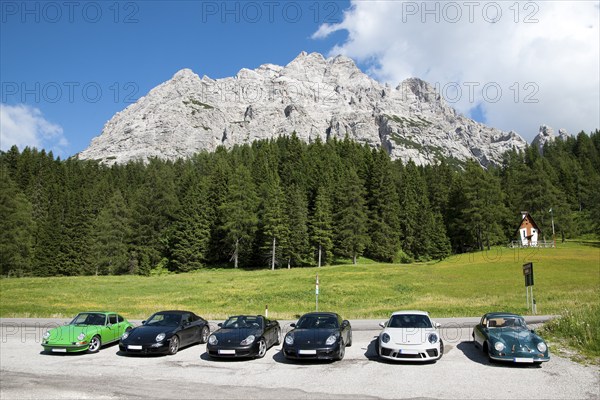 On the right Porsche 956 from the 50s from the left five Porsche sports car series 911 from different decades in front of alpine mountain scenery in Italian Alps Dolomites, Val di Zoldo, province Belluno, Italy, Europe