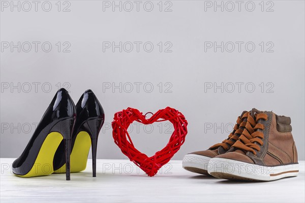 Red knitted heart with black womens high heel shoes and mens sneakers. Footwear on white background with copy space