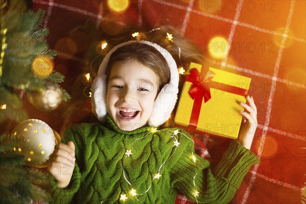 Girl in a warm knitted sweater and fur headphones is lying on a blanket near the Christmas tree with a gift box. Christmas Eve, a dream and a wish. New Year, festive atmosphere, garlands and bokeh