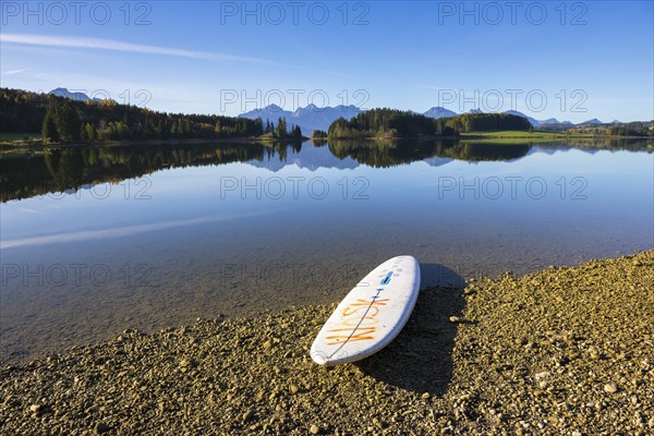 A calm lake with a surfboard on the shore, surrounded by mountains and a clear blue sky, Forggensee near Füssen, Ostallgäu, Bavaria, Germany, Europe