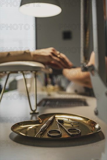 Tools for making nails in beauty parlor, scissors, cutter on a gold heart-shaped stand. Customer with cosmetologist in the background