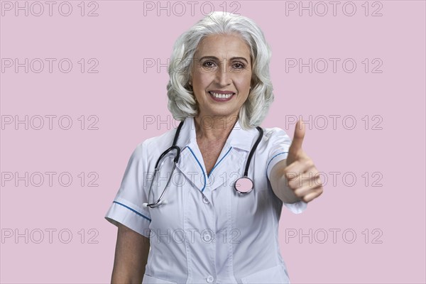 Successful senior woman doctor gesturing thumb up to camera. Joyful female doctor giving thumb up sign on color background