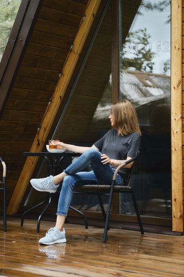 Woman sits in wicker chair near the table at wooden terrace of her forest cabin