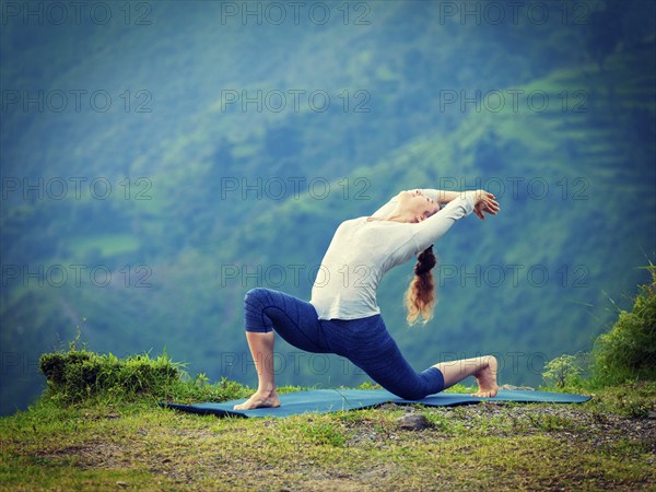 Yoga outdoors, sporty fit woman practices Hatha yoga asana Anjaneyasana, low crescent lunge pose posture outdoors in Himalayas mountains. Vintage retro effect filtered hipster style image