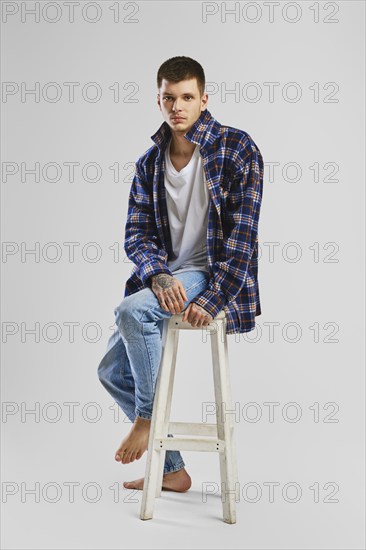 Full length studio portrait of young cocky man in shirt and jeans sitting on tall wooden chair