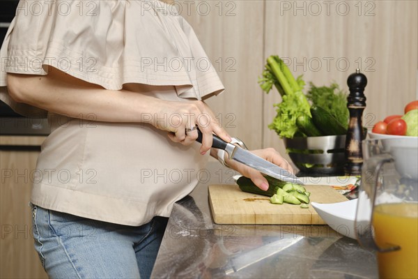 Closeup view of hands of pregnant unrecognizable woman in the kitchen cutting fresh and juicy cucumber for salad