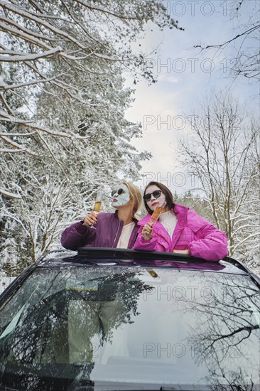 Two funny women with cosmetic sheet masks on their faces holding a glass of champagne leaning out of the car hatch