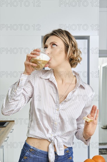 Young woman drinks cappuccino for breakfast at home, the plate with fried eggs and toasts stands on the table in front of her