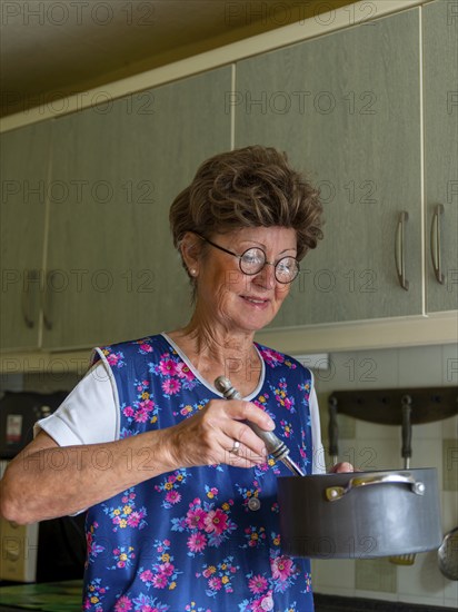 Granny with old smock apron, glasses and wig in the kitchen, holding cooking pot and ladle, Germany, Europe