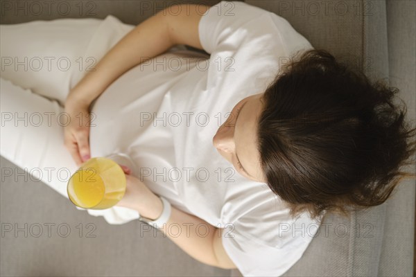 Top view of a pregnant woman sitting on the sofa with a glass of fresh orange juice in her hand