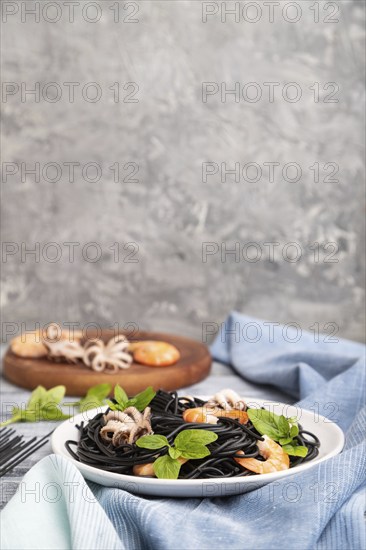 Black cuttlefish ink pasta with shrimps or prawns and small octopuses on gray wooden background and blue textile. Side view, copy space, selective focus