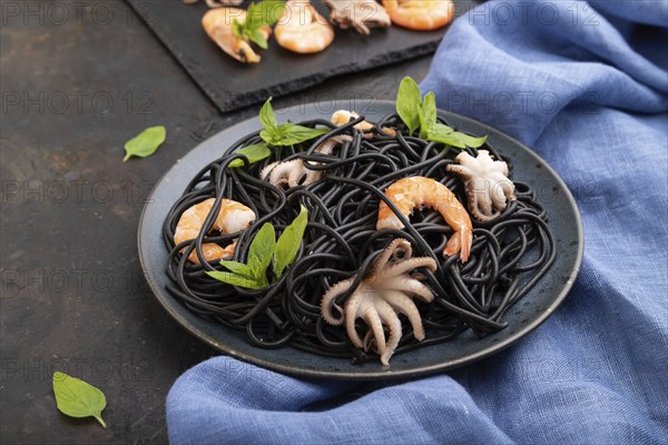 Black cuttlefish ink pasta with shrimps or prawns and small octopuses on black concrete background and blue textile. Side view, close up