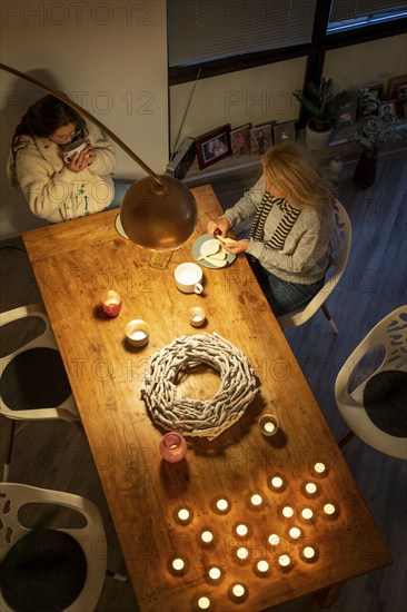 Symbolic image energy saving, cold winter, energy crisis, cold flat, mother and daughter dressed warmly at dinner, only one lamp on, candlelight, heating on to a minimum