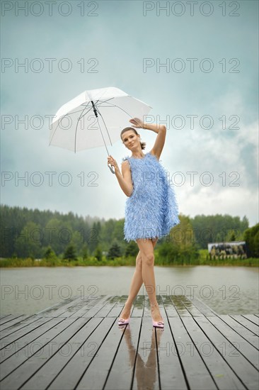 Stylish woman in blue mini dress and pink stilettos posing on a wooden dock with a transparent umbrella under the rain