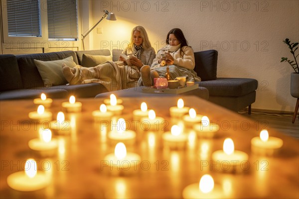 Symbolic image energy saving, cold winter, energy crisis, cold flat, mother and daughter lying on the sofa, warmly dressed, candlelight, heating on to a minimum