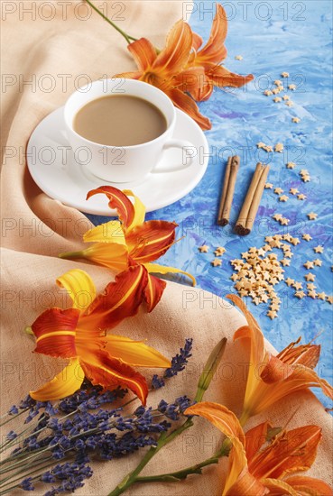 Orange day-lily and lavender flowers and a cup of coffee on a blue concrete background, with orange textile. Morninig, spring, fashion composition. side view