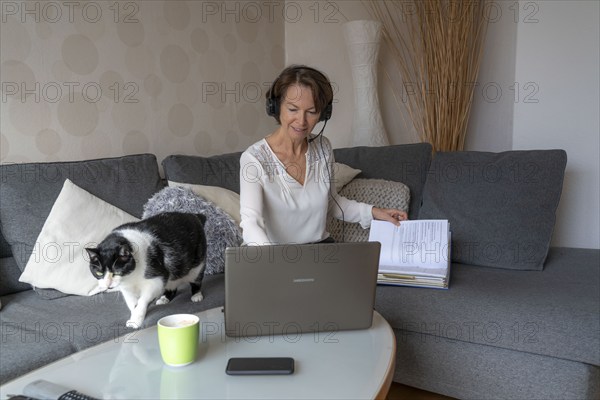 Woman, mid-50s, works from home, with laptop and communicates with colleagues via headset, home office, on the sofa