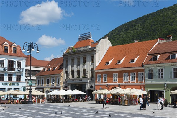 Sunny market square with historical buildings, people at street cafés and mountain in the background, town houses at the market square Piata Sfatului, Old Town, Brasov, Transylvania, Romania, Europe