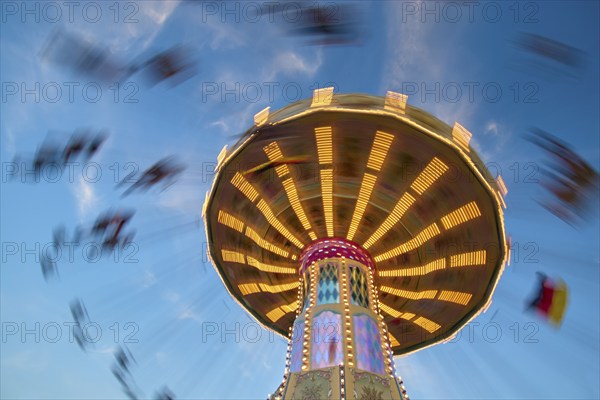 An illuminated carousel at dusk, spinning fast, in front of a blue sky with clouds and a Germany flag, Killiani Volksfest, Würzburg, Bavaria