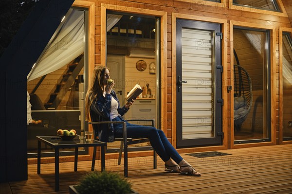 Side view of a woman relaxing on the terrace of modern chalet with a book and an apple in her hands