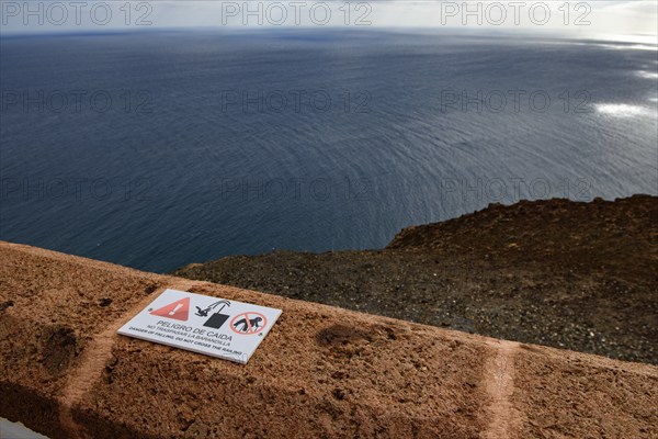 View from building complex of lighthouse Faro de la Entallada from 50s year 1953 1954 on 183 metres high rocks of cliff east coast of Fuerteventura on Atlantic Ocean, in the foreground on wall cornice warning sign warning of danger of falling into the depth, Las Playitas, Tuineje, Las Palmas, Fuerteventura, Canary Islands, Spain, Europe