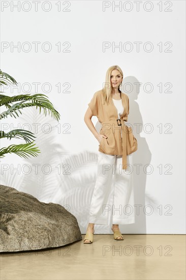 Trendy blonde woman in beige sleeveless top and loose white trousers posing under hard sunlight near the rock