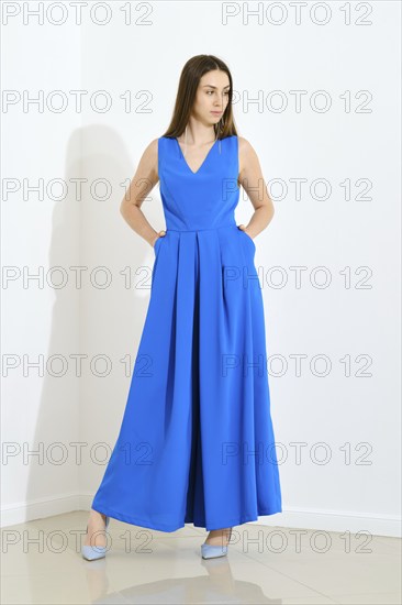 Full length portrait of stylish trendy woman in blue long sleeveless dress holding her hands in pockets