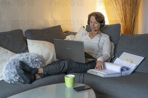 Woman, mid-50s, works from home, with laptop and communicates with colleagues via headset, home office, on the sofa