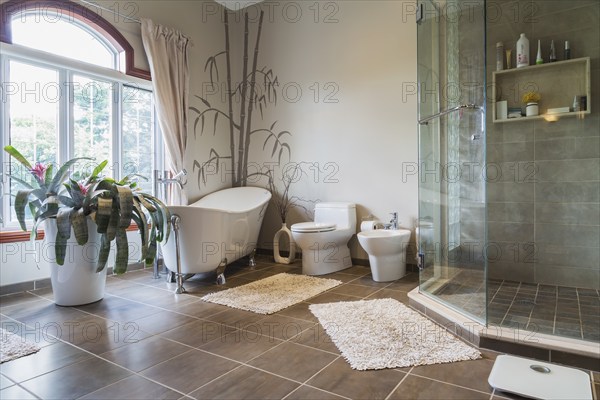 Freestanding bathtub, bidet, clear glass shower stall in en suite with grey nuanced ceramic tile floor on upstairs floor inside a luxurious residential home, Quebec, Canada, North America