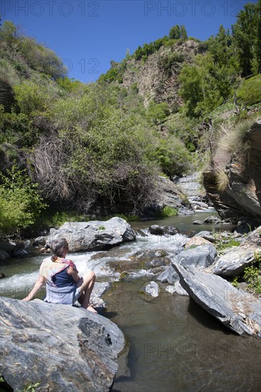 Woman sitting on rock by rapids in the channel of the River Rio Poqueira gorge valley, High Alpujarras, Sierra Nevada, Granada Province, Spain, Europe