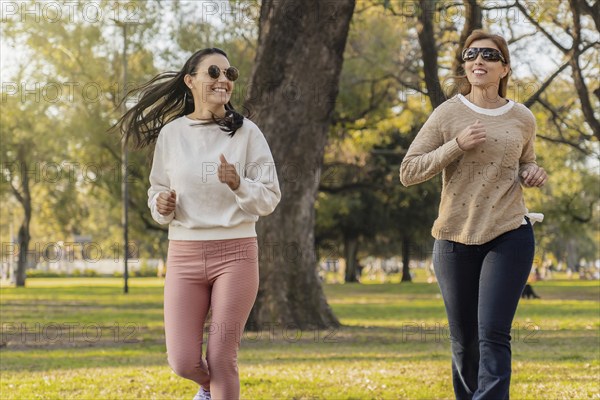 Two women are jogging side by side in a park, enjoying a sunny day outdoors. They smile as they exercise, dressed in casual athletic wear. The scene captures the joy of fitness and friendship