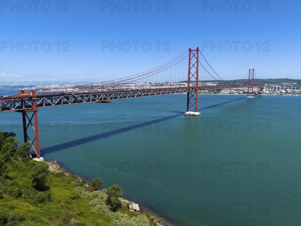 A large red bridge stretches across a wide expanse of water under a clear blue sky, surrounded by green countryside, aerial view, Ponte 25 de Abril, 25 April Bridge, suspension bridge over the Tagus, double-decker bridge, road and rail traffic, Lisbon, Lisboa, Portugal, Europe