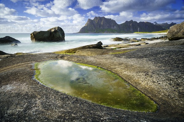 Seascape on the beach at Uttakleiv (Utakleiv), rocks and green seaweed in the foreground. In the background the mountain Hogskolmen. Good weather, blue sky with some clouds. Early summer. Long exposure. Uttakleiv, Vestvagoya, Lofoten, Norway, Europe