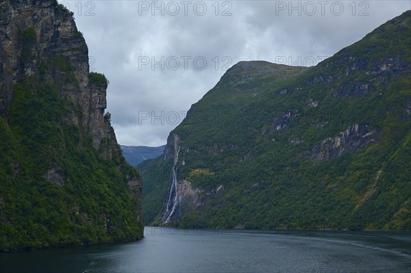 A fjord surrounded by rocky mountains under a cloudy sky with a waterfall in the background, Geiranger, Geiranger Fjord, Stranda, Romsdal, Norway, Europe