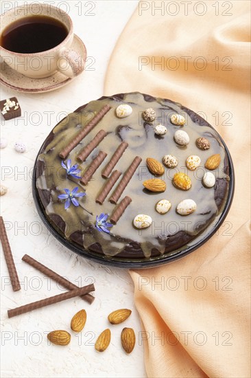 Homemade chocolate brownie cake with caramel cream and almonds with cup of coffee on a white concrete background and orange textile. Side view, close up