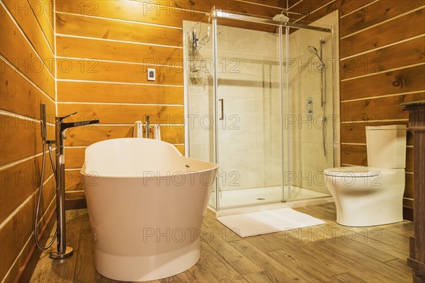 Freestanding bathtub with high tank toilet and clear glass shower stall in en suite with imitation wood ceramic tile flooring inside a milled Eastern white pine timber and flat log profile home, Quebec, Canada, North America