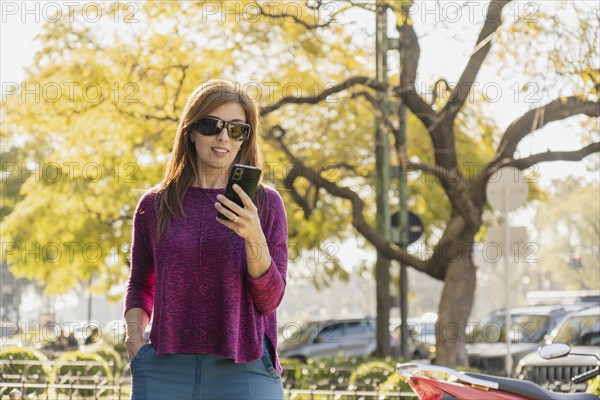 Stylish middle-aged woman smiles as she looks at her smartphone in a park at sunset. Wearing sunglasses and a purple sweater, she keeps one hand in her pocket, enjoying the relaxed outdoor setting