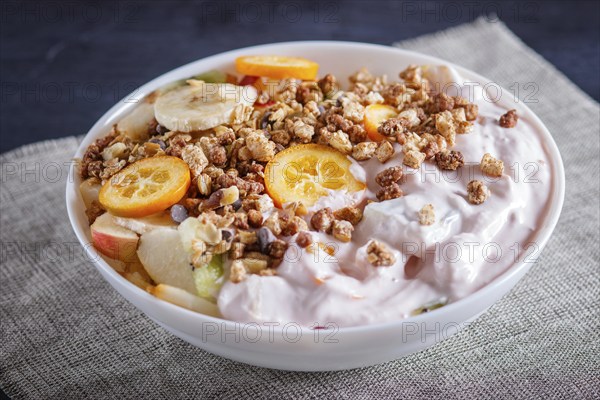 Vegetarian salad of bananas, apples, pears, kumquats, kiwi with granola and yogurt on black woodem background, close up, selective focus