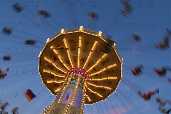 An illuminated carousel at dusk, spinning fast, in front of a blue sky with clouds and a Germany flag, Killiani Volksfest, Würzburg, Bavaria