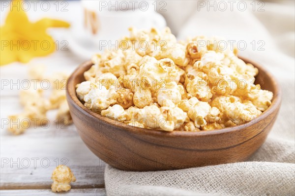 Popcorn with caramel in wooden bowl and a cup of coffee on a white wooden background and linen textile. Side view, close up, selective focus