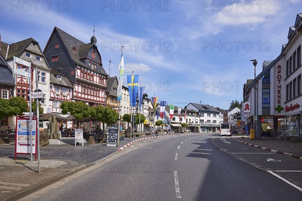 Main street with bus stop Adenau Markt, buildings, half-timbered houses, flags and trees under blue sky with cirrus clouds in Adenau, Eifel, Landkreis Ahrweiler, Rhineland-Palatinate, Germany, Europe