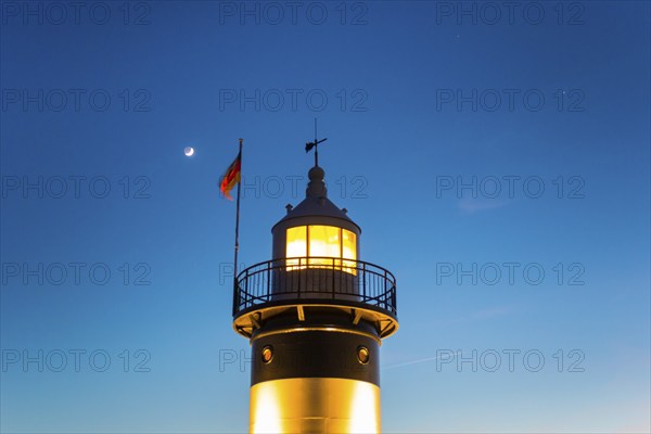 Upper segment of an illuminated lighthouse at night, black and white lighthouse, called 'Kleiner Preuße', with a clear blue sky and a rising waxing moon, German flag waving in the wind, bright beacon, twilight, evening, dusk, sea in the background, maritime, calm atmosphere, North Sea resort Wremen, Lower Saxony Wadden Sea National Park, World Heritage Site, Wurster North Sea coast, Land Wursten, district Cuxhaven, Lower Saxony, Germany, Europe