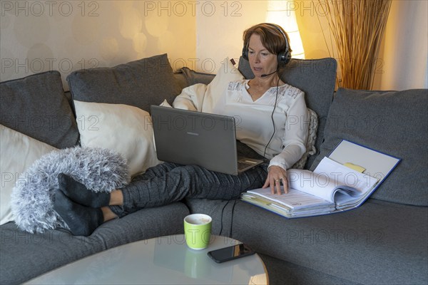 Woman, mid-50s, works from home, with laptop and communicates with colleagues via headset, home office, on the sofa