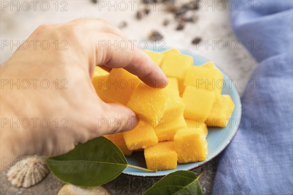 Dried and candied mango cubes on blue plate with hand on brown concrete background and linen textile. Side view, close up, selective focus, vegan, vegetarian food concept