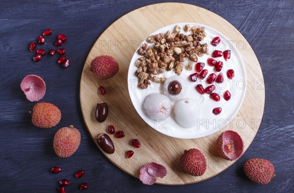 Greek yogurt with lychee, pomegranate seeds and granola in a white plate on a black wooden background, top view, flat lay, close up