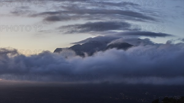 Mystical scene of a mountain peak shrouded in clouds at dusk with lights of a town in the valley, Taygetos Mountains, Mani Peninsula, Peloponnese, Greece, Europe