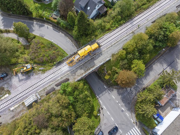 A yellow construction machine on railway tracks crosses a bridge on a winding road in a green neighbourhood, tamping machine, Hermann Hessebahn, Althengstett, Black Forest, Germany, Europe