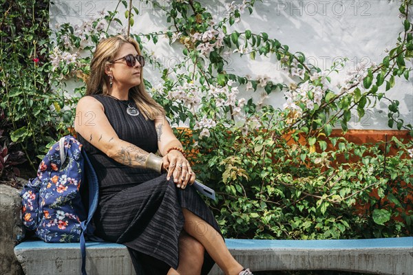 A fashionable woman with tattoos and a flower backpack sits on a bench, surrounded by vibrant flowers and lush greenery. She exudes a relaxed and calm air in an outdoor setting.